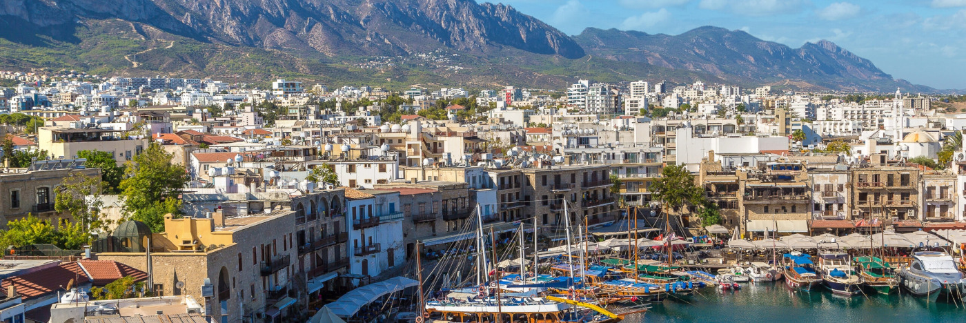 A panoramic shot of a harbour in North Cyprus with colourful saiing boats docket. Behind are the small white buildings making up the harbourside and town while in the background are gentle mountains dotted with trees and green.