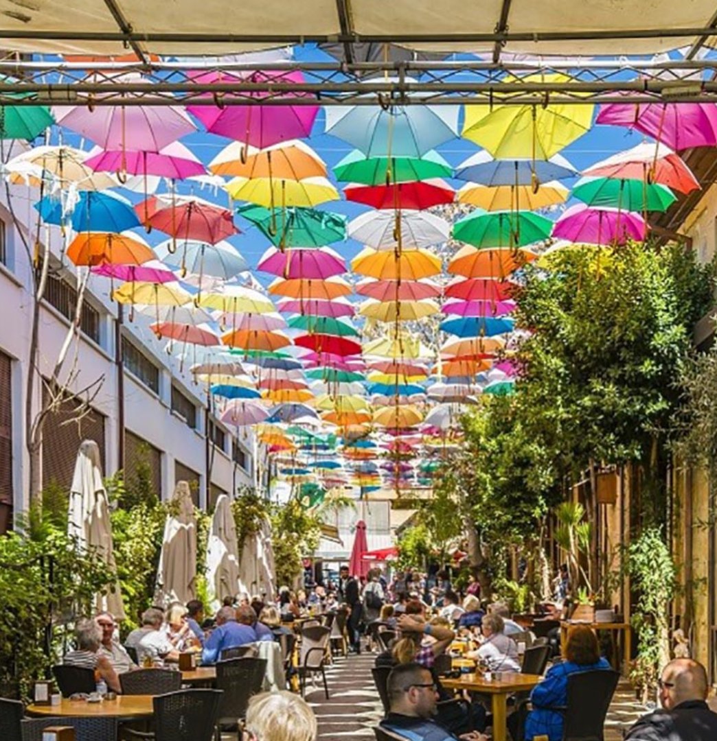 A sunny colonnade in Nicosia where numerous people are sitting around tables eating and drinking. Hanging above are a multiple brightly coloured umbrellas suspended on wires.